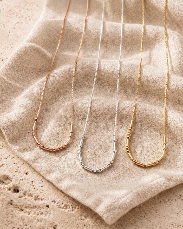 Blancora Gold, Silver and Rose Gold - delicate necklace pendants in rose gold, silver, and yellow gold, laid out on a textured beige fabric surface.