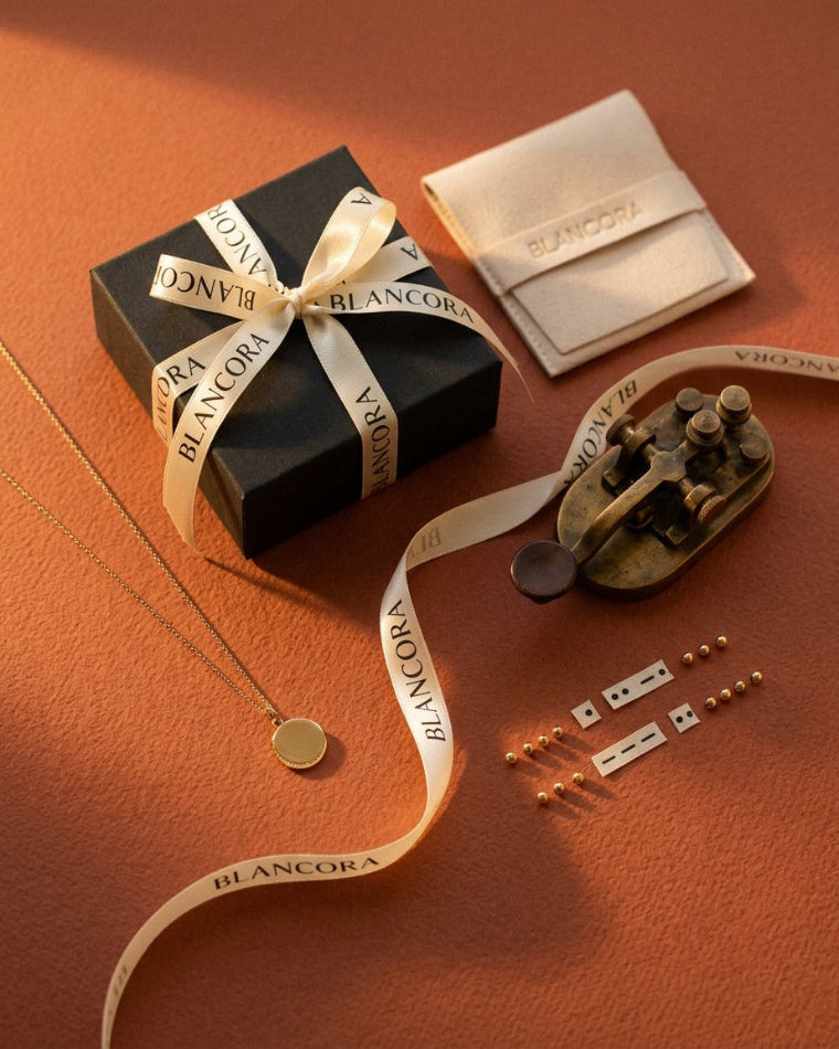 Blancora jewelry gift set arranged on a warm terracotta background, including a black gift box with a cream ribbon printed with the BLANCORA name, a cream fabric pouch, a gold pendant necklace, a vintage-style gold clasp, small gold beads, and metal findings.