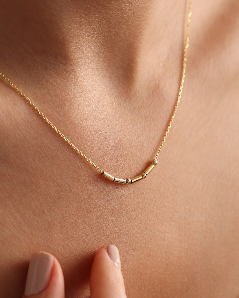 A delicate morse code gold chain necklace with three small cylindrical beads at the center, worn on a person's neck.